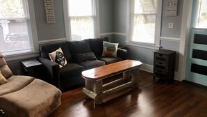 A living room with a dark-colored sofa, a wooden coffee table, and a beige armchair. There are two windows with blinds, and a door with a blue frame. - The Apollo (Charleston)
