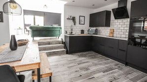 A modern kitchen with dark cabinetry, a wooden dining table, and a green sofa in the adjacent living area. - Ty Coch (Ruthin)
