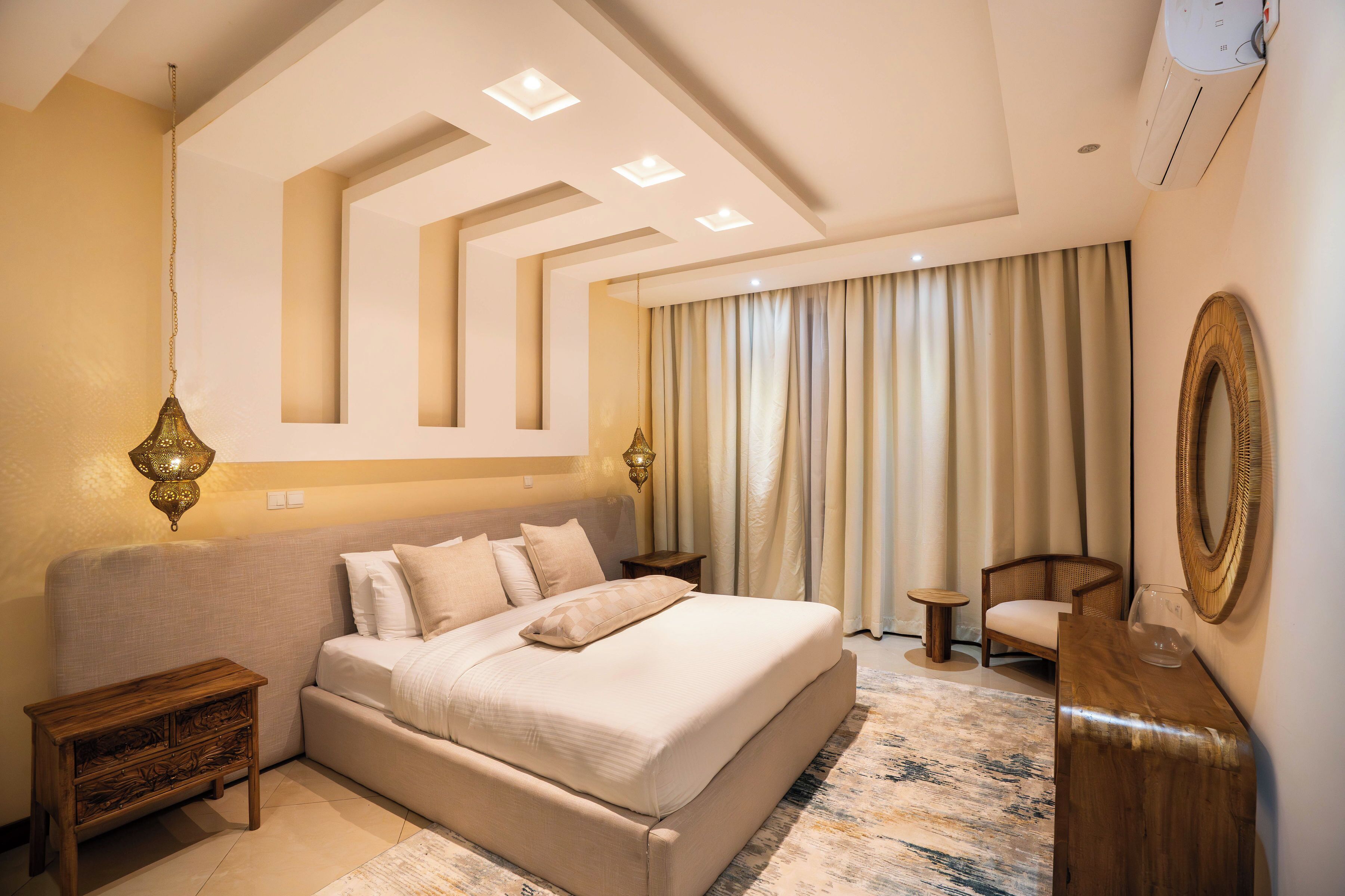 A modern hotel room with a large bed, a wooden nightstand, a small side table, a chair, and a decorative mirror.