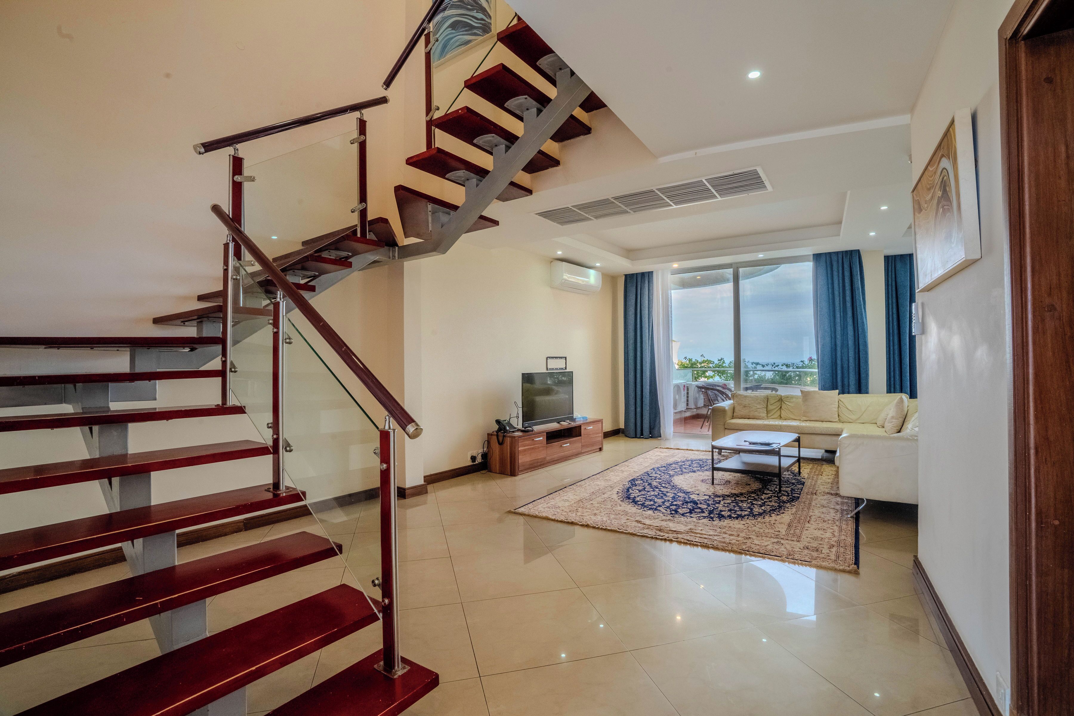 A modern living room with a staircase, a sofa set, a flat-screen TV, and a large window with curtains.