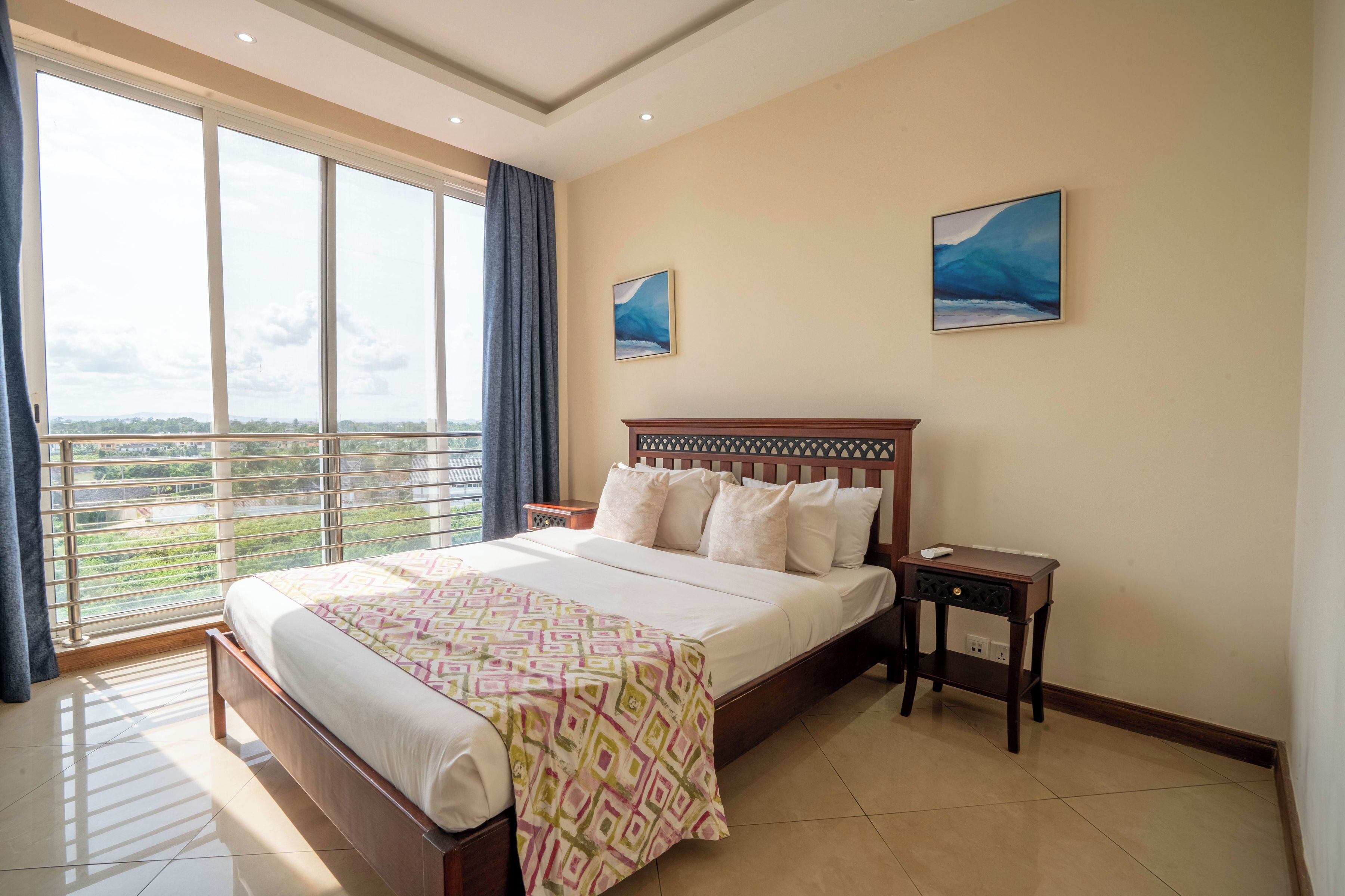Habitación de hotel con una cama grande, una mesita de noche y vistas de la ciudad a través de amplias ventanas.