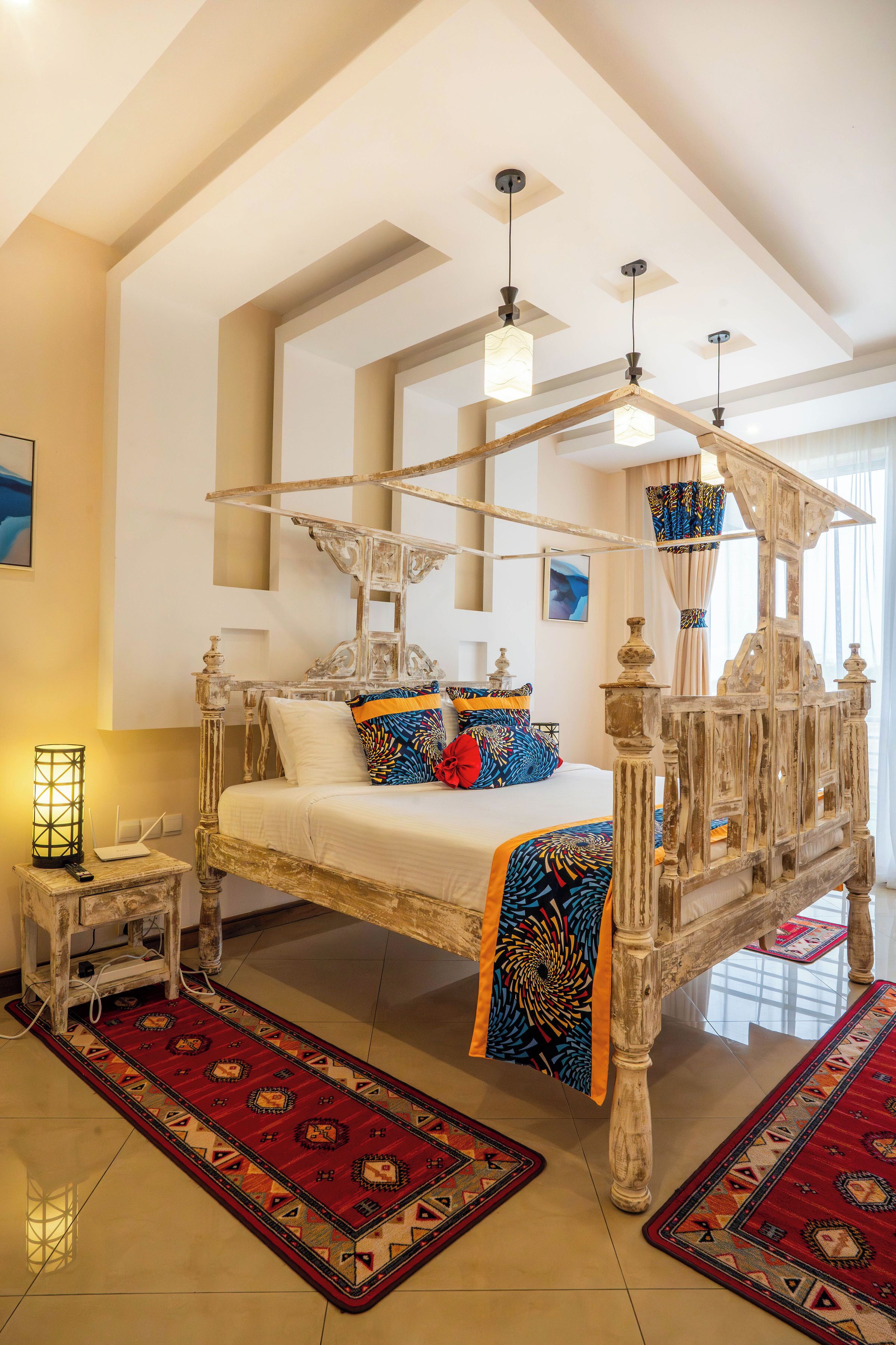 A four-poster bed with a wooden frame and ornate carvings, a bedside table with a lamp, and a red patterned rug on the floor.