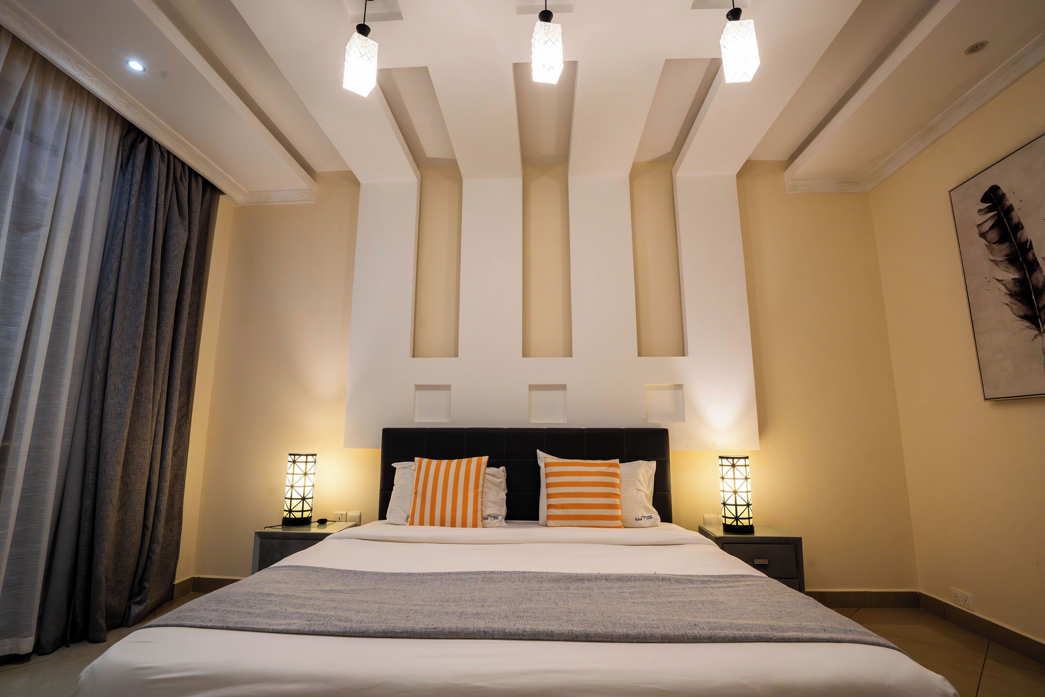 A modern hotel room with a large bed, two bedside tables with lamps, a headboard with built-in lighting, and a large window with curtains.