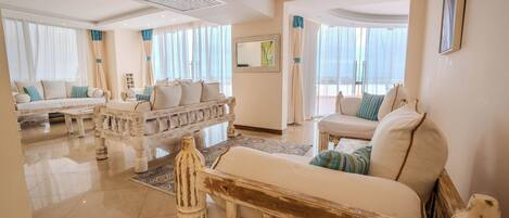 A spacious living room with a white sofa set, a wooden bench, and a large window with sheer curtains.