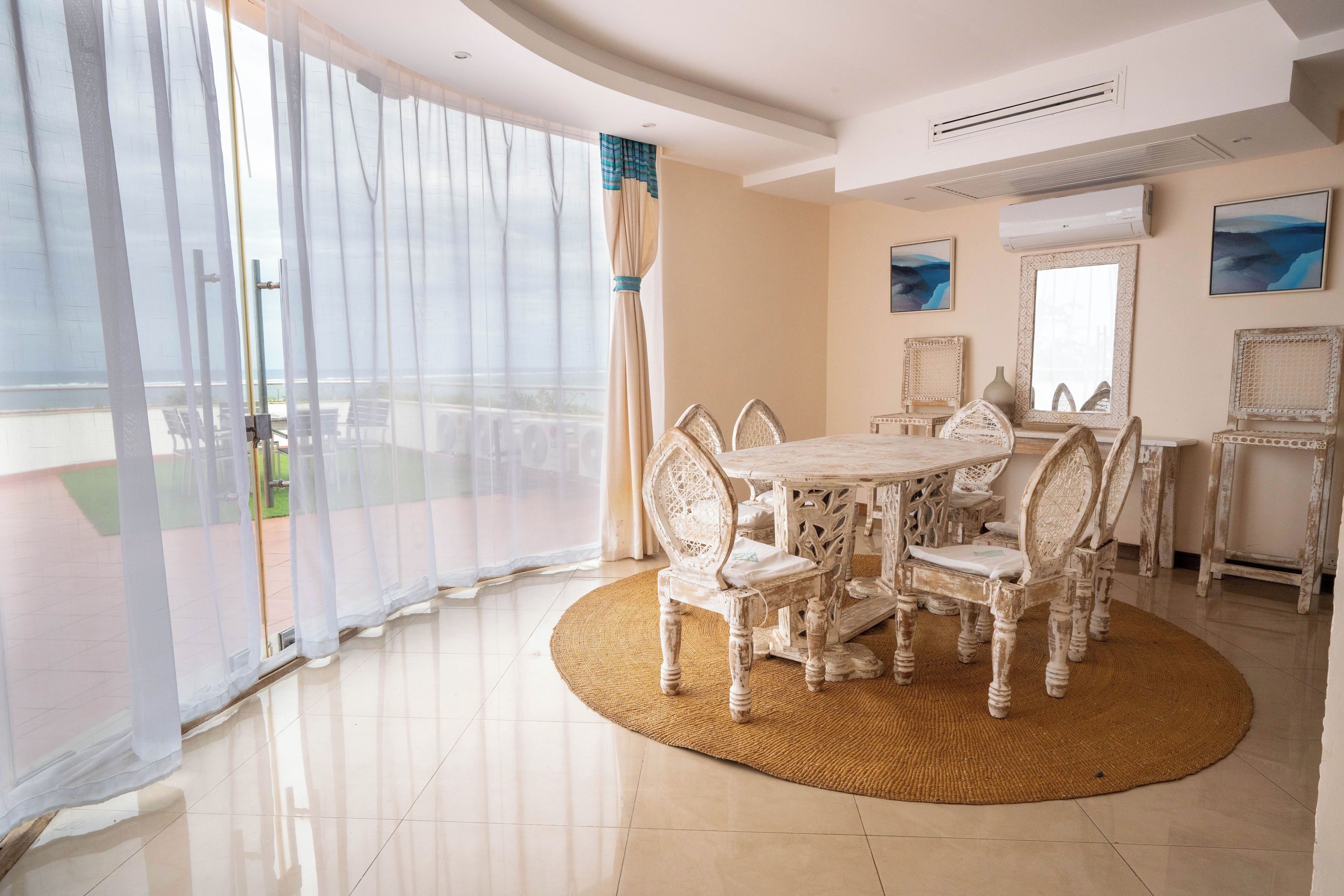 Una habitación amplia con un gran ventanal corredera que da a un balcón, una mesa de comedor redonda con sillas ornamentadas y un espejo grande en la pared.