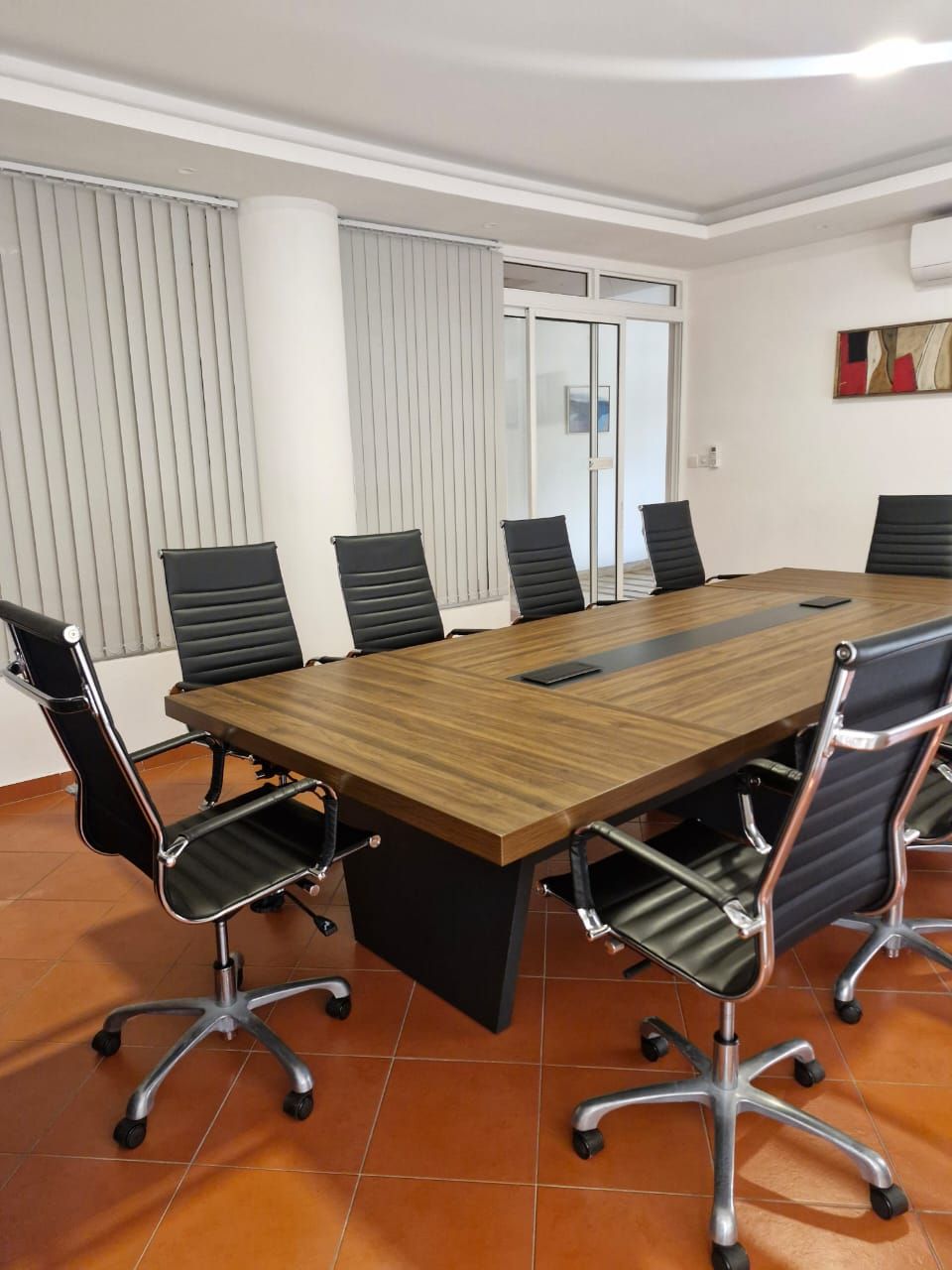 A conference room with a large wooden table, black leather chairs, and a glass partition.