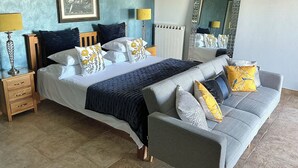 Un dormitorio con cama, sofá, mesita de noche, lámpara, espejo y un cuadro en la pared.