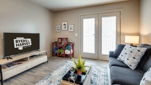 A living room with a flat-screen TV, a sofa, a coffee table with a tray, a potted plant, a wooden side table, and a door with a window. - The Lofts of Downtown (540 N 2nd St, #211) (Clarksville)