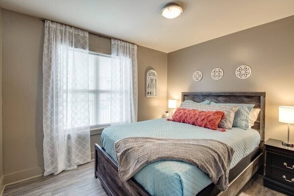 Un dormitorio con cama, mesita de noche, ventana con cortinas traslúcidas y detalles decorativos en la pared.