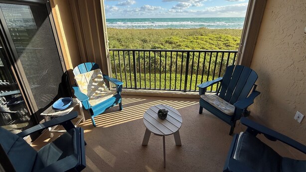A balcony with two blue chairs, a small round table, and a view of the ocean and beach. - Ocean view oasis (Jensen Beach)