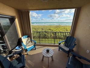 Un balcón con dos sillas azules, una mesa redonda pequeña y vistas al océano y la playa.