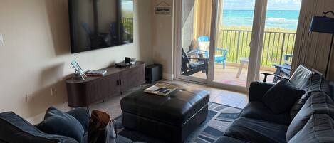 Un salon doté d’un grand téléviseur à écran plat, d’un canapé de couleur sombre, d’une table basse et d’une porte-fenêtre coulissante donnant sur un balcon avec vue sur l’océan.