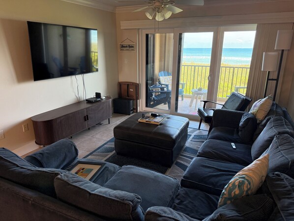 Una sala de estar con un televisor de pantalla plana grande, un sofá, una mesa de centro y un ventanal corredizo que da a un balcón con vistas a la playa.