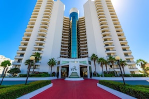 Een hoog hotel met een centrale blauwe toren, meerdere balkons en een rode pad dat naar de ingang leidt.