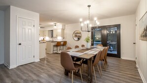 A dining area with a wooden table and chairs, a kitchen in the background, and a large window with a view.