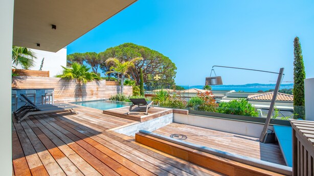 Um deck de madeira com piscina, cadeiras de descanso e vista para o mar.