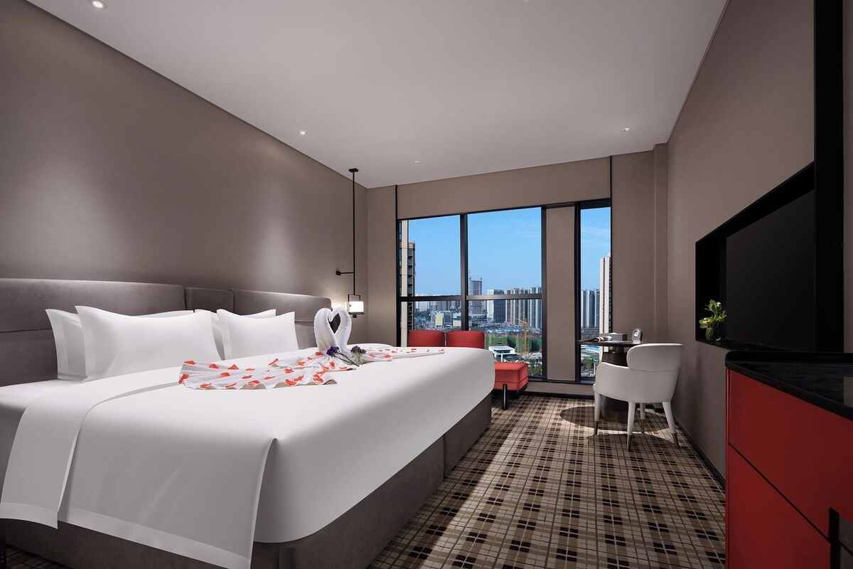 A modern hotel room with a large bed, a desk, a TV, and a city view.