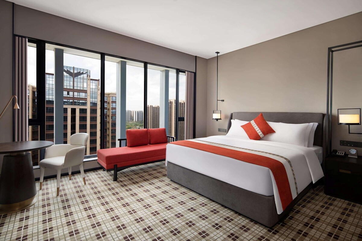 A hotel room with a large bed, a seating area with a red cushion, a desk, and a view of buildings through large windows.
