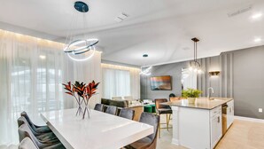 Uma cozinha moderna com ilha central, área de jantar com mesa e cadeiras brancas e sala de estar com sofá e mesa de centro.
