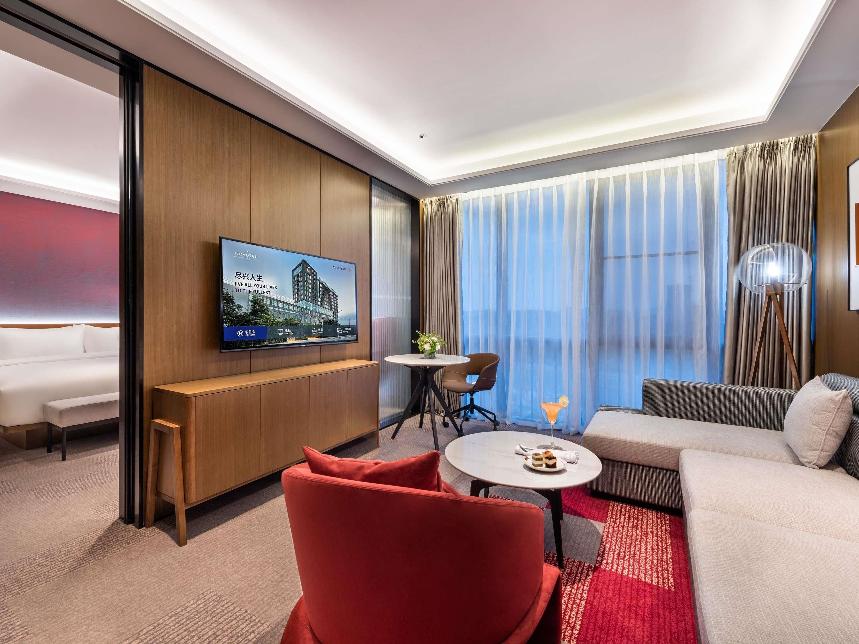 A hotel room with a bed, a sofa, a red armchair, a small table with a drink, a TV mounted on the wall, and a window with curtains.