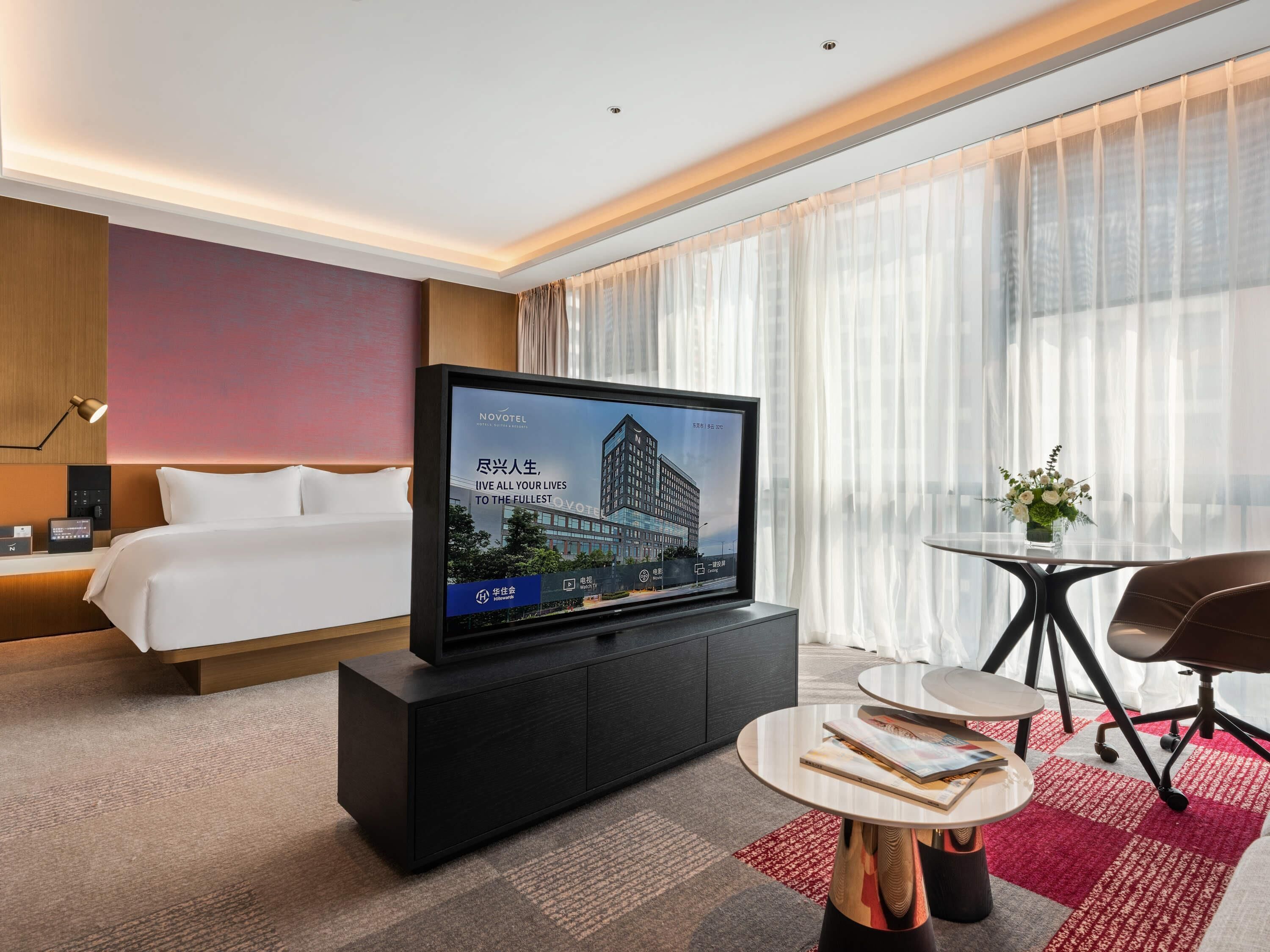 A modern hotel room with a bed, a flat-screen TV on a cabinet, a small round table, and a chair.