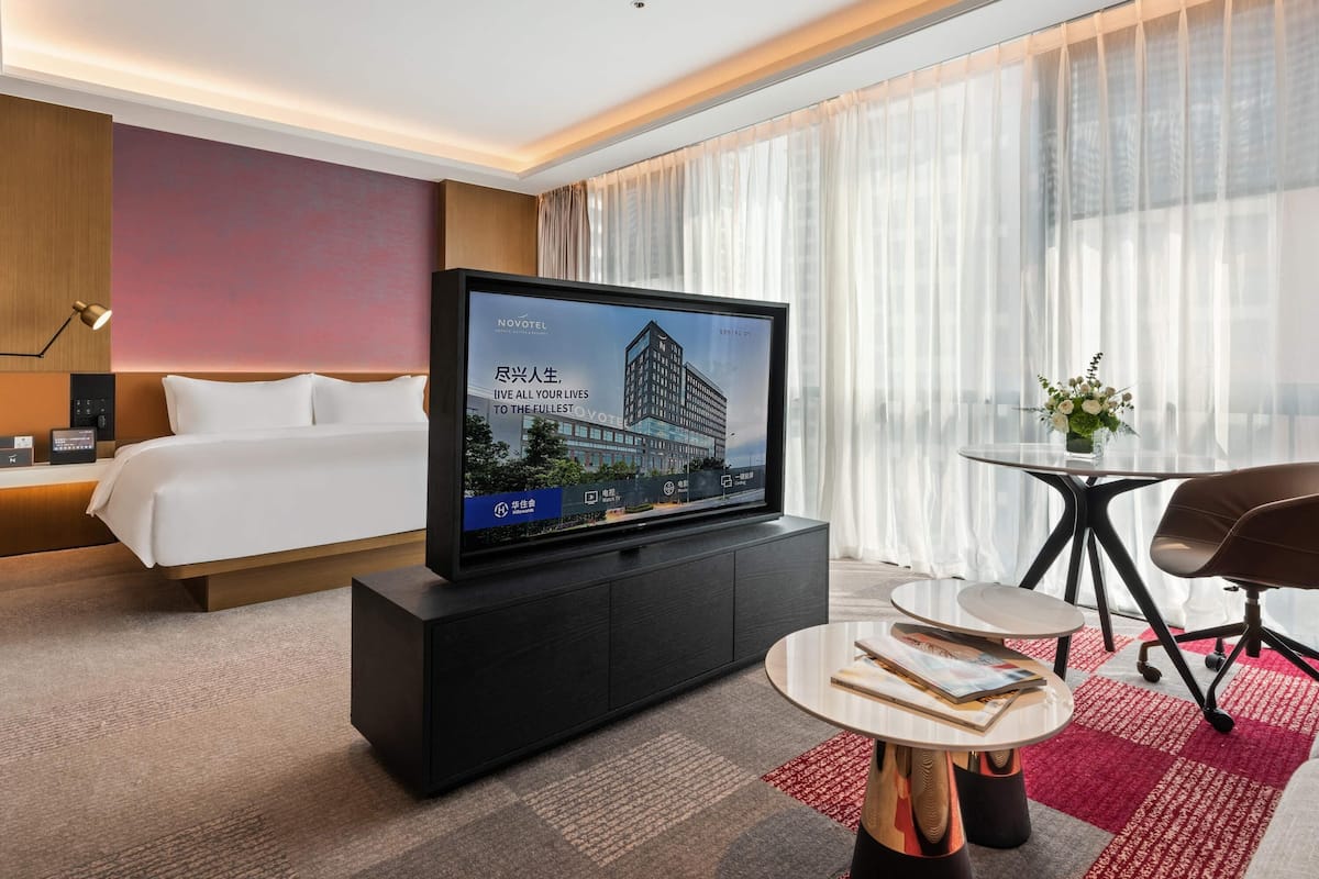 A modern hotel room with a bed, a flat-screen TV on a cabinet, a small round table, and a chair.
