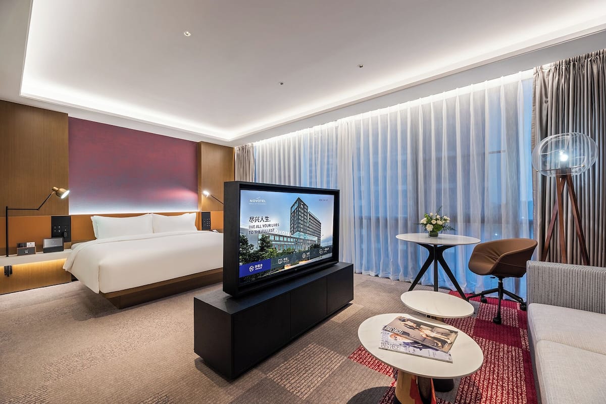 A modern hotel room with a bed, a TV on a black stand, a small round table with a magazine, a chair, and a large window with curtains.