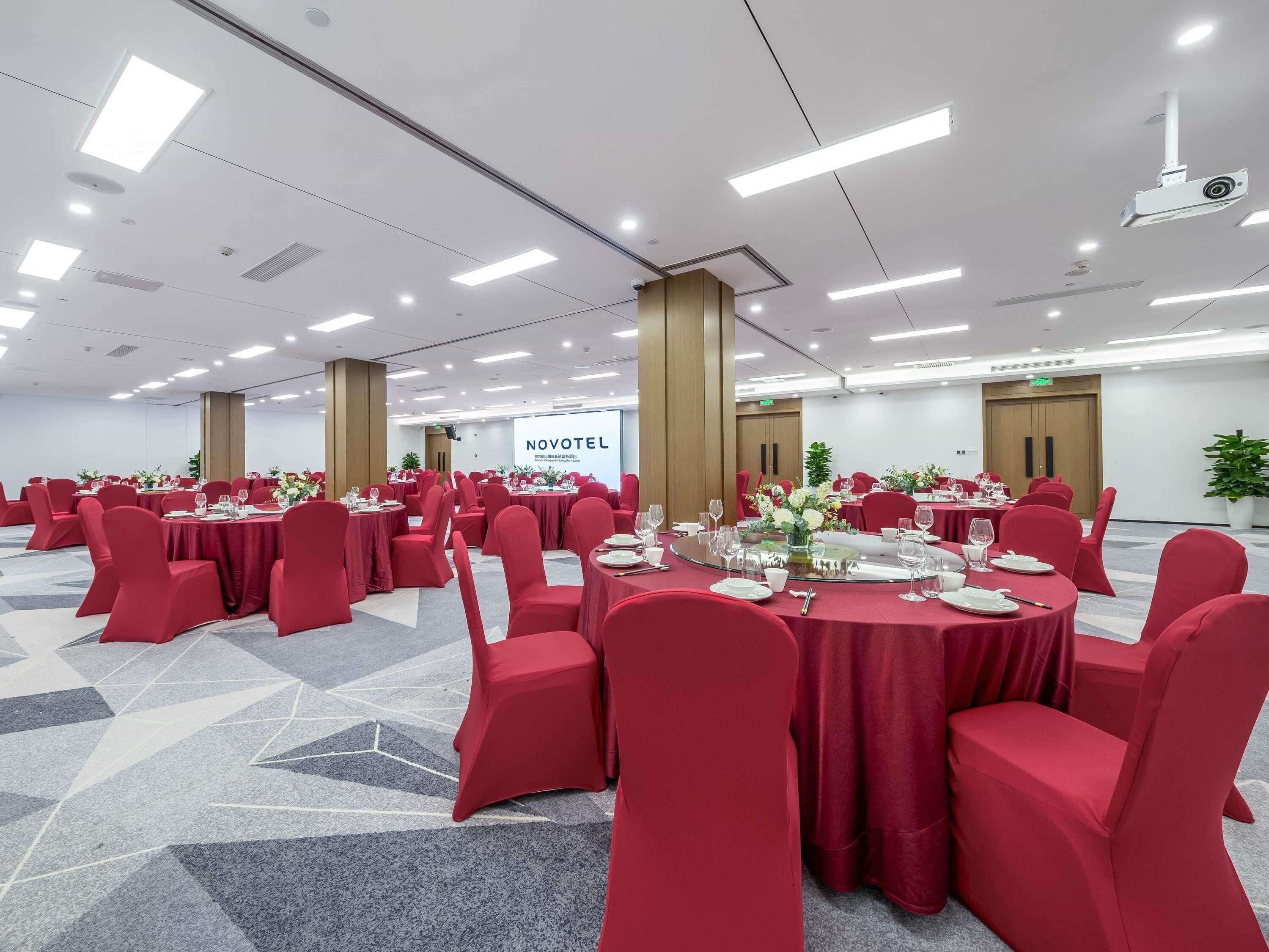 Une salle de conférence avec des tables rondes, des chaises rouges et une enseigne « NOVOTEL ».