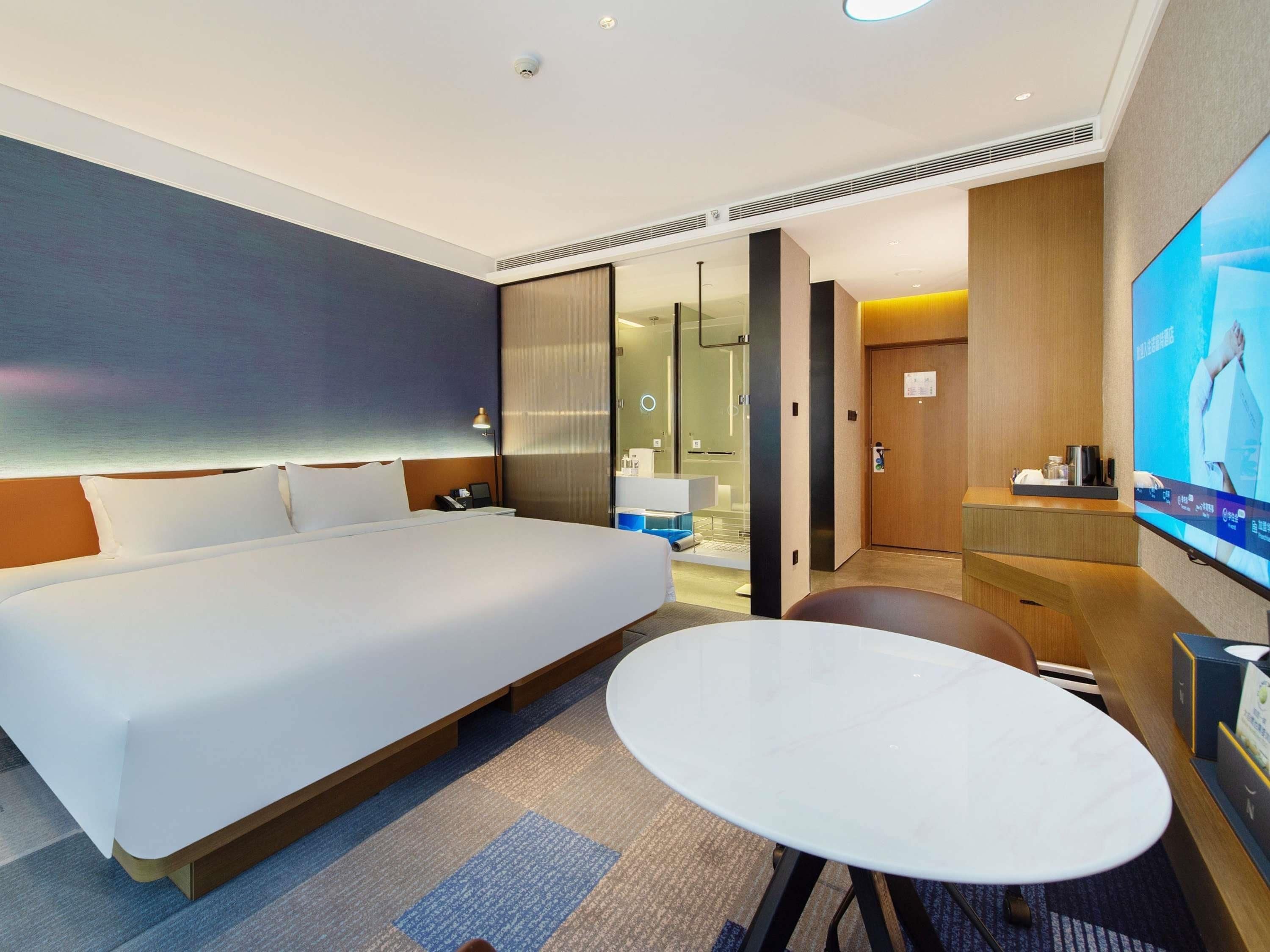 Una habitación de hotel moderna con una cama grande, una mesa redonda, un televisor y un baño visible a través de una puerta de vidrio.