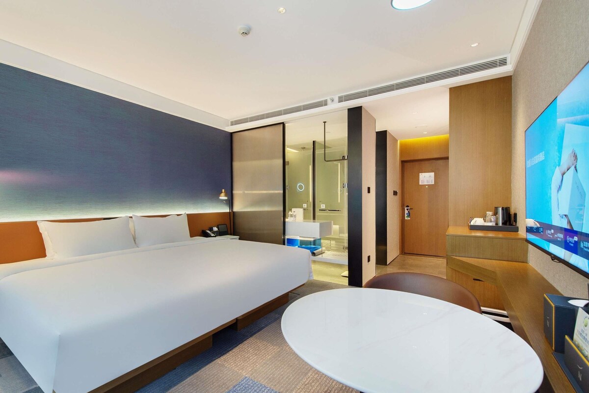 Una habitación de hotel moderna con una cama grande, una mesa redonda, un televisor y un baño visible a través de una puerta de vidrio.