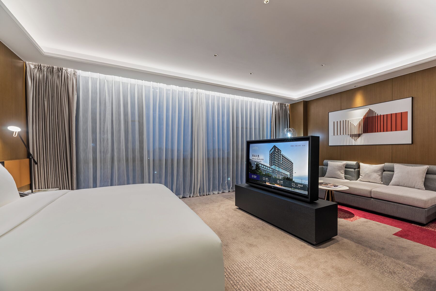 Quarto de hotel moderno com cama grande, televisão de tela plana, sofá e uma janela ampla com cortinas.