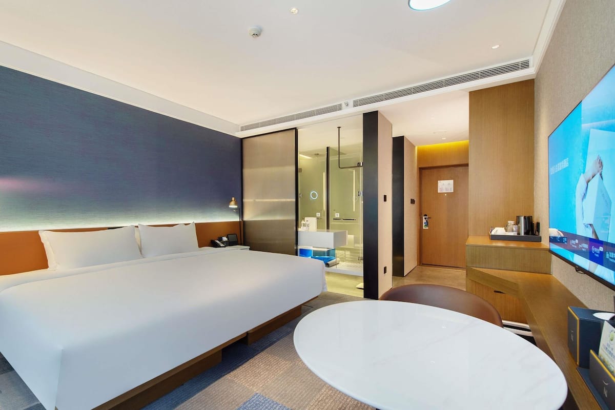 A modern hotel room with a large bed, a round table, a TV, and a bathroom visible through a glass door.