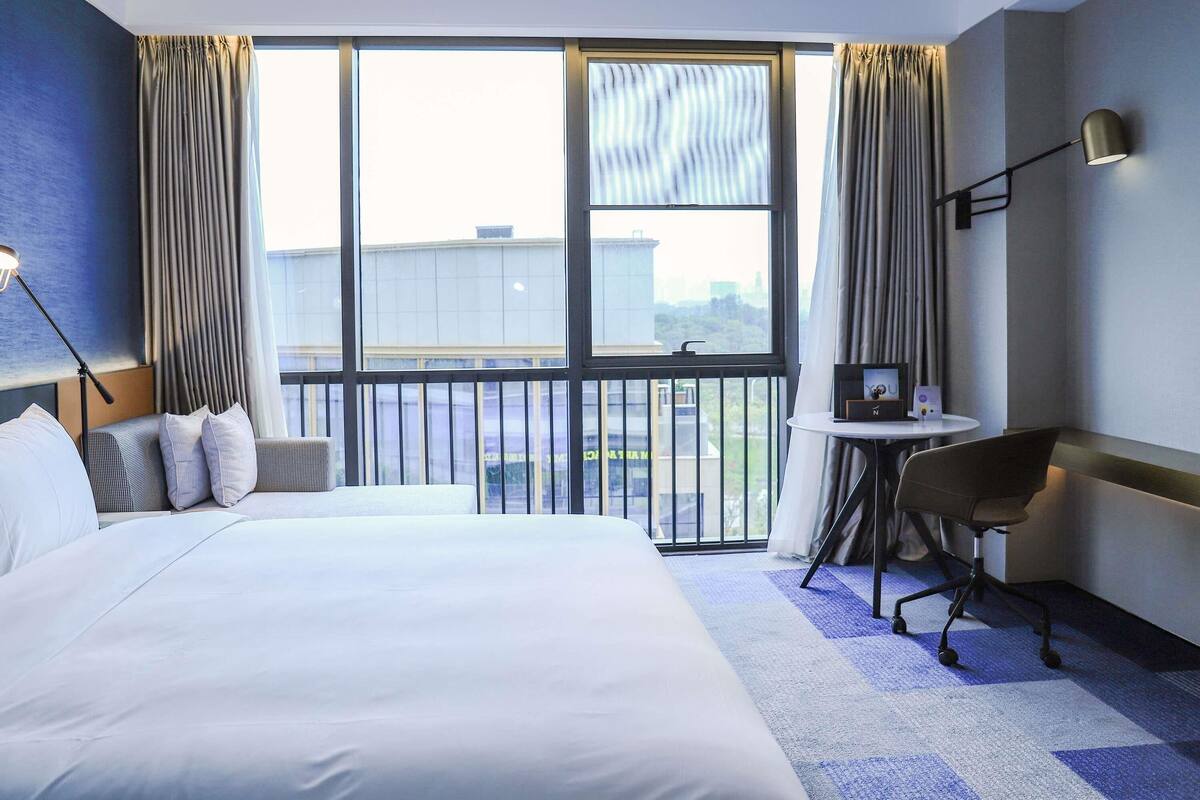 A hotel room with a large bed, a desk, a chair, and a view of the city through large windows.