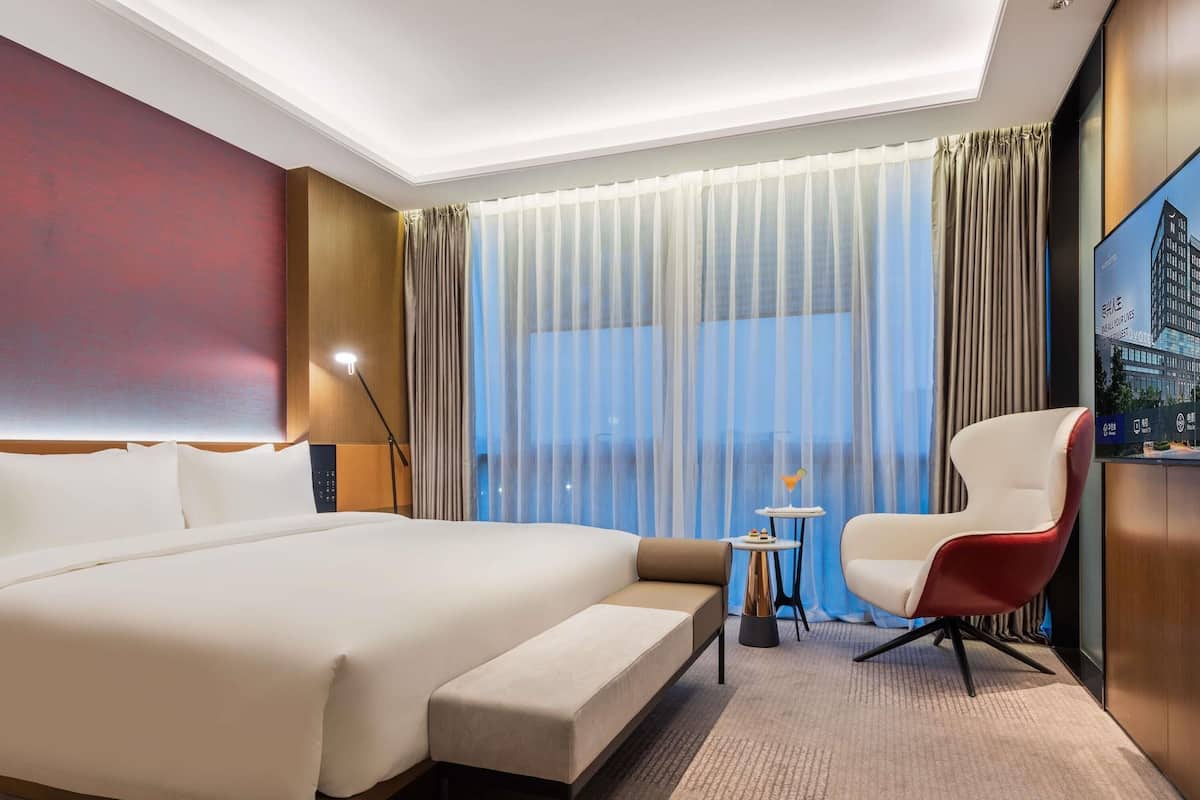 A hotel room with a large bed, a small table with a drink, a red chair, and a TV displaying a building.