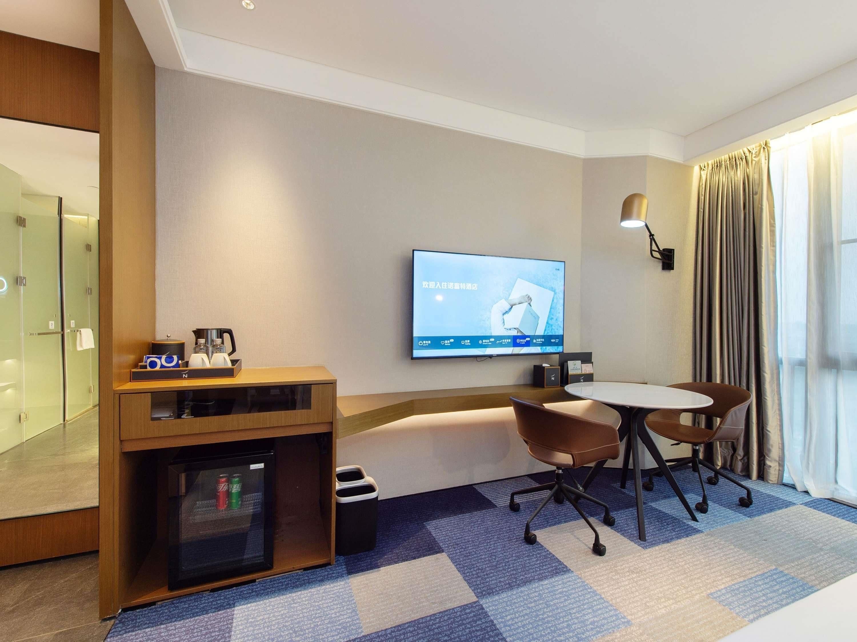 A hotel room with a flat-screen TV mounted on the wall, a small refrigerator, a coffee maker, a trash bin, and a seating area with two chairs and a table.