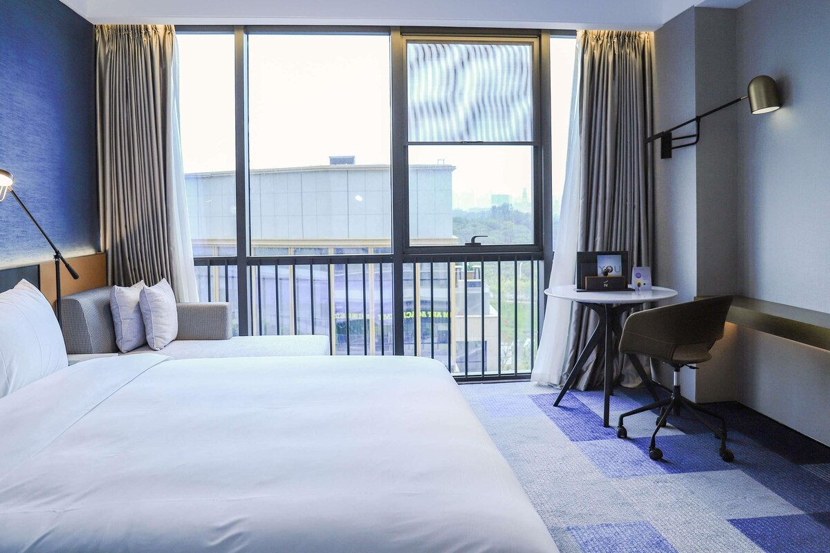 Habitación de hotel con una cama grande, un escritorio, una silla y vistas a la ciudad a través de amplios ventanales.