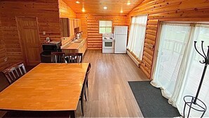 Interieur van een houten blokhut met een houten eettafel, stoelen, een keuken met een fornuis en koelkast, en een raam met gordijnen.