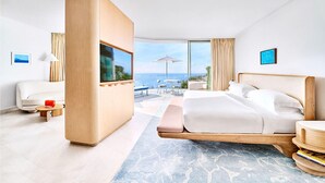 A modern hotel room with a large bed, a TV, a seating area with a table and chairs, and a view of the sea.