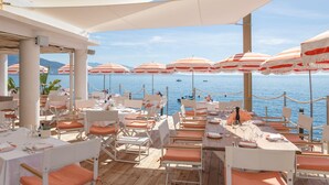 An outdoor dining area with tables and chairs, umbrellas, and a view of the sea.
