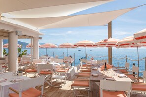 Area pranzo all'aperto con tavoli e sedie, ombrelloni e vista sul mare.