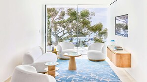 A modern living room with a large glass door leading to an outdoor area, featuring white sofas, a round coffee table, and a blue patterned rug. - The Maybourne Riviera (Roquebrune-Cap-Martin)