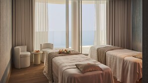 A hotel room with two beds, a sitting area with a chair, a small table, and a view of the sea.