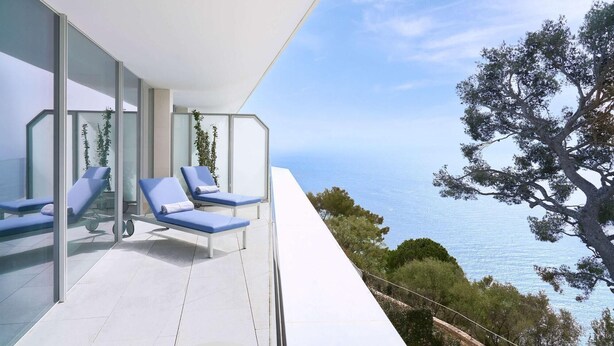A modern outdoor lounge area with glass walls and blue lounge chairs, overlooking a coastal landscape with trees and the sea. - The Maybourne Riviera (Roquebrune-Cap-Martin)