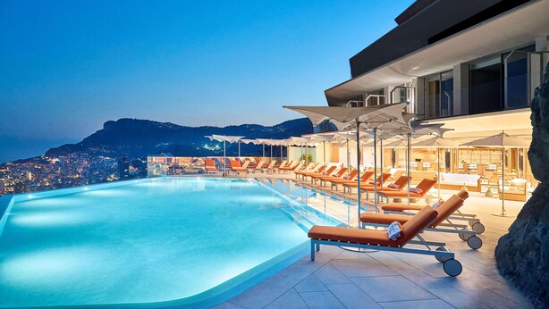 A pool area with lounge chairs and umbrellas, overlooking a cityscape at dusk.