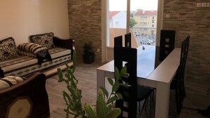 A living room with a sofa, a dining table, chairs, and a large window with a view of buildings outside. - Beautiful modern apartment near the sea (Frontignan)