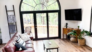 A spacious living room with a leather sofa, a wooden coffee table, and a black wicker chair. A large arched window offers a view of a wooden deck and trees.