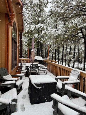 A wooden deck with snow-covered chairs and tables, surrounded by snow-laden trees.