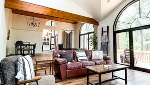 A spacious living area with a leather sofa, a wooden coffee table, and a dining table. There are large windows providing natural light and a view of the outdoors.