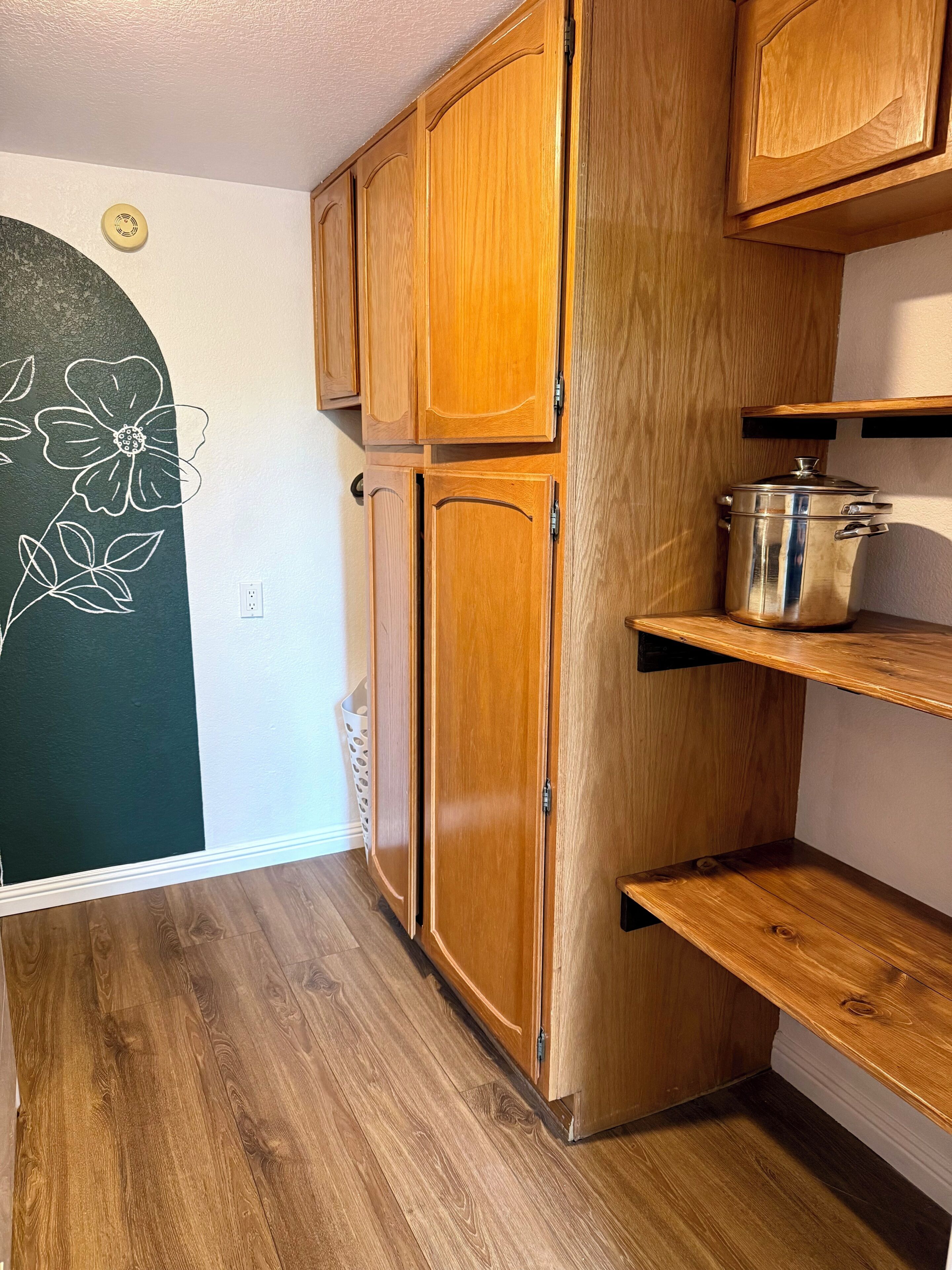 Une cuisine avec des meubles de rangement en bois, une casserole en acier inoxydable et un mur orné d’un motif floral.