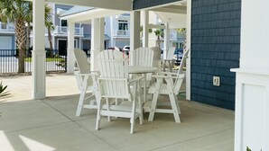 A covered patio area with white Adirondack chairs and a table, a blue exterior wall, and a view of neighboring buildings. - Beach Getaway: Stunning, Family-Friendly w/ Pool. Walk to Shopping + Dining (Atlantic Beach)